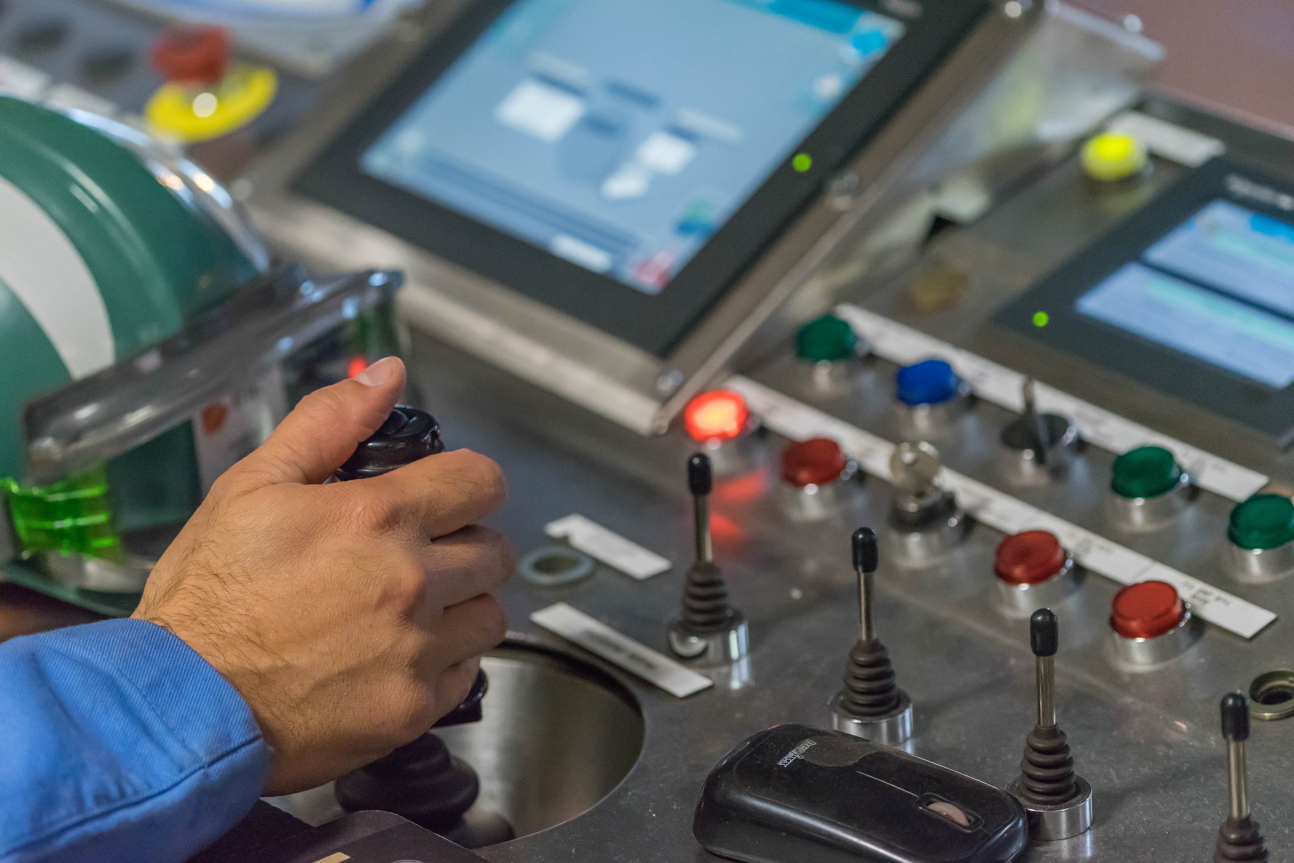 Operator hand on a joystick at a control desk with pushbuttons and HMI screens