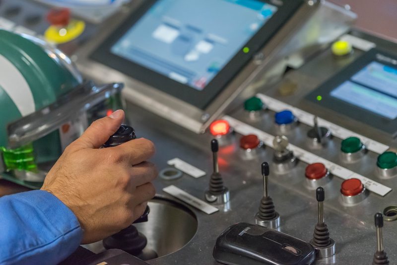 Operator hand on a joystick at a control desk with illuminated pushbuttons and HMI screens